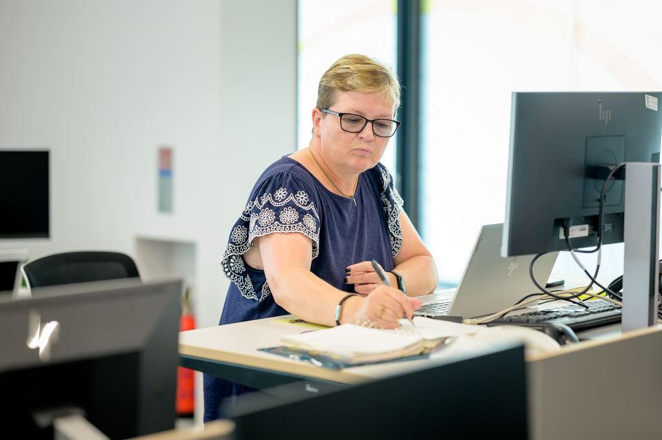 A woman at an office desk working from a notebook beside a laptop and monitor