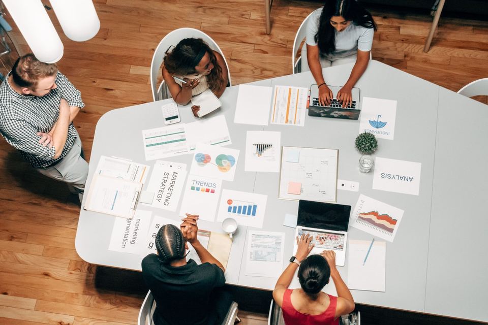 Five colleagues around a table covered in printed marketing charts and strategy notes, seen from above