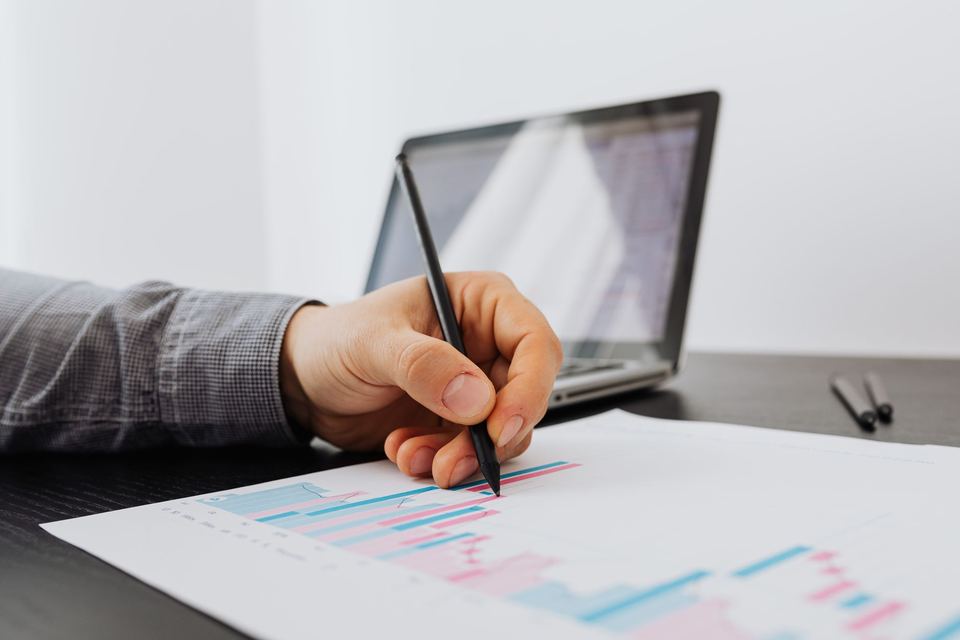 A hand holding a pen over a printed bar chart, with a laptop behind